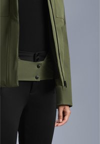 Olivno zelena jakna na zadrgo s prilegajočim krojem, ki ima pritisne gumbe na pasu in je v kompletu s črnimi ozkimi hlačami.