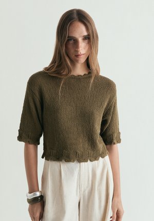 Jeune femme aux longs cheveux bruns porte un pull marron texturé à manches courtes et un pantalon crème taille haute sur un fond clair.