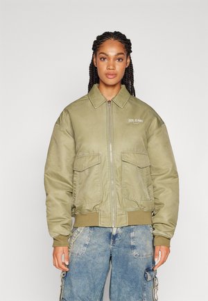 Kvinne med flettet hår iført en lyse olivengrønn zip-up jakke og løse blå denim cargo-bukser mot en ensfarget bakgrunn.