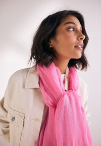 Hellrosa Schal aus weichem, luftigem Stoff, um den Hals gebunden, getragen mit einer beigen Jacke; glatte Textur und fließendes Design.