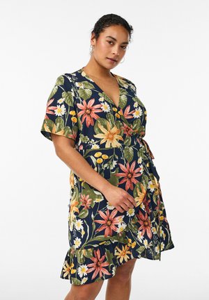 Moteris dėvi tamsiai mėlyną susiaučiamą suknelę su dideliais rožinės, geltonos ir baltos spalvų gėlių raštais, trumpomis rankovėmis ir raukšlėtu apvadu ant baltos fono.