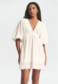 Weißes Baumwollkleid mit tiefem V-Ausschnitt, Puffärmeln, gerafftem Bund und Spitzenbesatz am Saum. Besitzt gestickte Details überall.