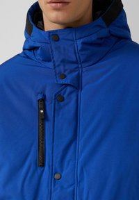 Blaue Jacke mit einer Reißverschlusstasche, Druckknöpfen und hohem Kragen. Glatte Textur mit matter Oberfläche und schwarzen Akzenten am Kragen.