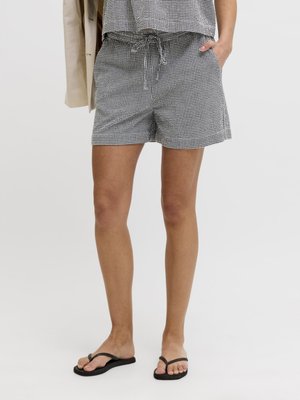 Nő fekete-fehér kockás, zsinóros rövidnadrágban és fekete papucsban, egyik kezét a zsebében tartja, bézs dzsekit a karján hord.