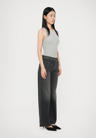 Ženska stoji oblečena v sivo brezrokavno majico, visoko pasove svetleče črne široke hlače in črne špicaste čevlje pred enobarvno ozadje.