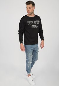 Czarny sweatshirt z napisem "TOP GUN", jasnoniebieskie zniszczone dżinsy i białe sneakersy, z okrągłym dekoltem i dopasowanymi rękawami.