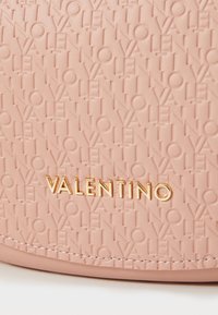 Στρογγυλή ροζ δερμάτινη τσάντα με ανάγλυφο μοτίβο "Valentino", που διαθέτει χρυσό λογότυπο μπροστά και ορατές ραφές κατά μήκος των άκρων.