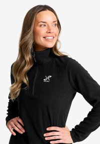 Leende kvinna med långt hår som bär en svart fleece med kvartzip och RVRC-logotyp, händerna på höften, ser åt sidan mot vit bakgrund.