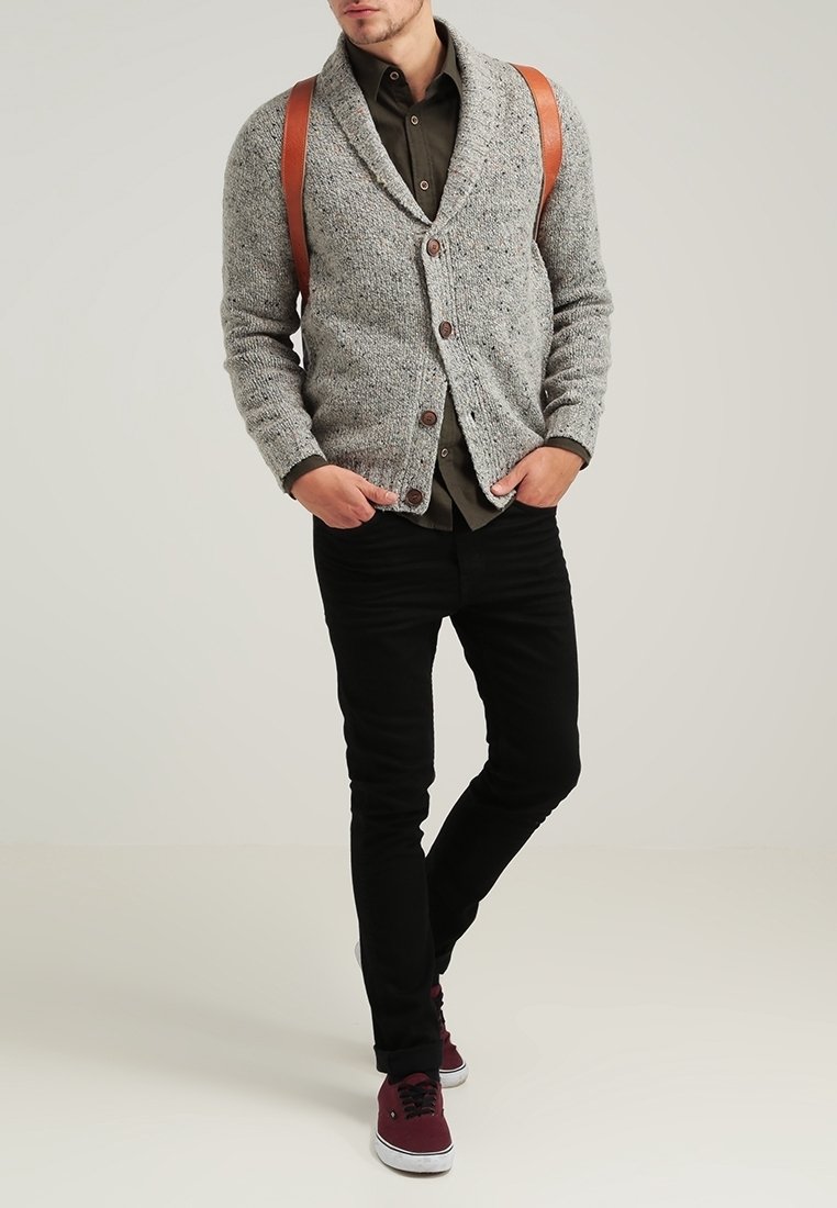 Homme portant un cardigan gris boutonné, une chemise olive, un pantalon noir, des baskets bordeaux et des bretelles marron, debout les mains dans les poches.