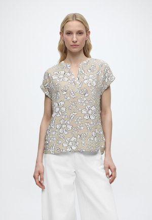 Femme aux longs cheveux blonds portant un chemisier beige à manches courtes avec un motif floral et un pantalon large blanc, debout devant un fond blanc uni.