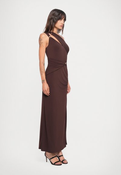 Lang, bruin, getailleerd jurkje met asymmetrische halslijn, gedrapeerd materiaal bij de taille en een gouden accentdetail. G gedragen met zwarte hakken met bandjes.