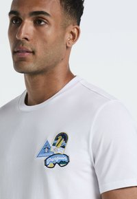 Witte katoenen t-shirt met geborduurde patches van een pinguïn, bergen en een trofee, met een ronde halslijn en korte mouwen.