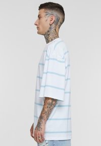 Tricou alb din bumbac cu dungi orizontale de culoare albastru deschis, mâneci scurte, croială lejeră și guler tip crew. Tatuaje notabile pe brațe și gât.