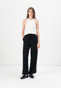 Zwarte wijde broek met een trekkoord in de taille, gecombineerd met een aansluitend wit geribbeld tanktop. Model staat tegen een effen witte achtergrond.