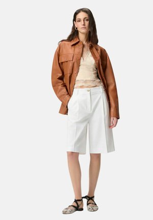 Femme portant une veste en cuir marron, un top en dentelle crème, un short blanc au genou et des chaussures plates à motifs avec des brides noires, debout devant un fond blanc.