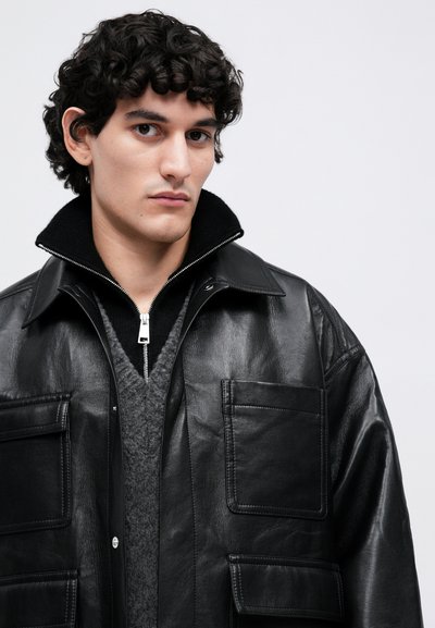 Giovane uomo con capelli ricci scuri indossa una giacca di pelle nera sopra un maglione scuro con zip e un gilet grigio strutturato, guardando avanti.