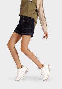 Sneakers bianche con una finitura testurizzata, dettagli accentuati trasparenti e una suola spessa. Abbinati a pantaloncini di jeans scuri e una maglietta verde oliva.