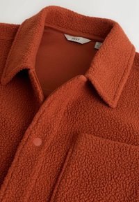 Jopica iz rjave flis tkanine z teksturirano površino, z zapenjanjem na gumbe in ovratnikom. Vsebuje prsni žep in gladko notranjo podlogo.