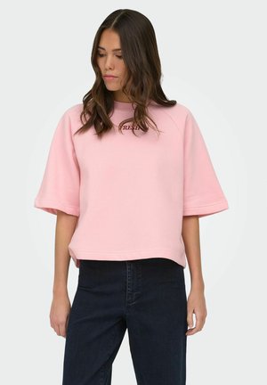 Femme portant un sweat-shirt rose clair à manches courtes et amples avec un petit texte sur la poitrine, et un jean noir taille haute, debout devant un fond uni.