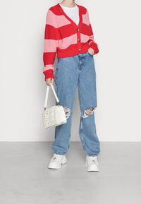 Rood- en roze gestreepte cardigan met een V-hals en knoopsluiting, gedragen over een wit T-shirt, gecombineerd met lichtblauwe gescheurde jeans en een witte handtas.