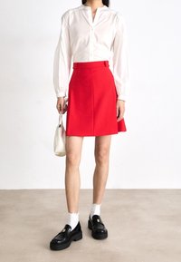 Jupe rouge trapèze avec boutons latéraux, associée à une chemise blanche à manches longues. Les accessoires comprennent un petit sac blanc et des mocassins noirs.