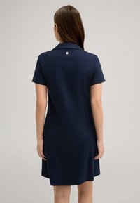Marineblauwe polojurk met een kraag, korte mouwen en een knoopdetail op de rug, gemaakt van een gladde stof met een licht uitlopende silhoeet.