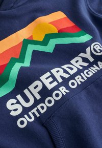 Sudadera azul marino con el texto "SUPERDRY" en blanco, diseño de montaña colorido con acentos en naranja, amarillo y verde, hecha de tela suave.