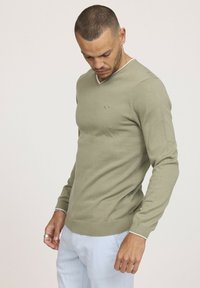 Pull vert clair en tricot doux, avec un col en V, des manches longues, des poignets et un ourlet côtelés, et un petit logo sur la poitrine.