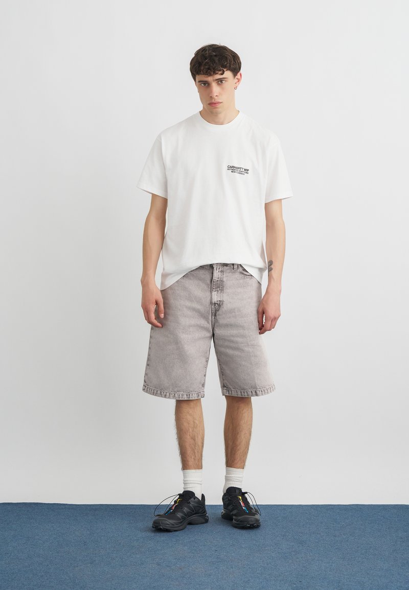 Giovane uomo in piedi che indossa una t-shirt bianca con grafica, pantaloncini grigi in denim, calzini bianchi e sneakers nere su un tappeto blu contro una parete bianca.