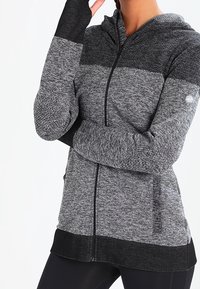 Grå og sort lynlåstrøje med blød tekstur, med farveblokdesign, tommelhuller og justerbar snøre på hætten.