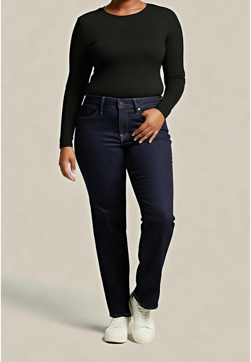 Femme portant un haut noir à manches longues, un jean bleu foncé et des baskets blanches, debout devant un fond uni.