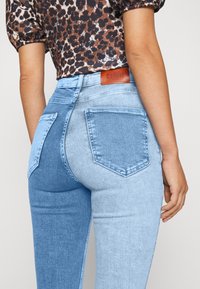 Ljusblå denimjeans med ett lapptäckesdesign, som har fickor, blekta sektioner och en läderetikett vid midjan som säger "DENIM."