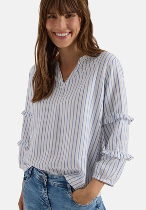 Femme souriante portant un chemisier blanc à fines rayures verticales bleues et manches à volants, associée à un jean bleu en denim.