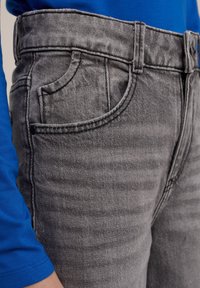 Graue Denim-Jeans mit geradem Schnitt, ausgestattet mit einer Vordertasche, Gürtelschlaufen und leicht gewaschener Textur. Kombiniert mit einem blauen Langarmoberteil.