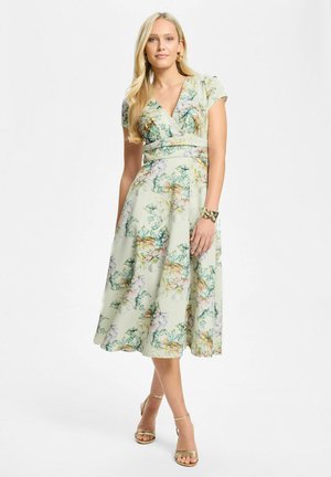 Femme aux longs cheveux blonds portant une robe midi verte pâle à fleurs avec des manches courtes, des sandales et un bracelet dorés.
