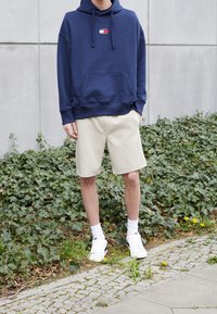 Felpa cappuccio blu navy con tasca frontale e logo, abbinata a pantaloni corti beige e sneakers bianche con dettagli neri. Foliage verde sullo sfondo.