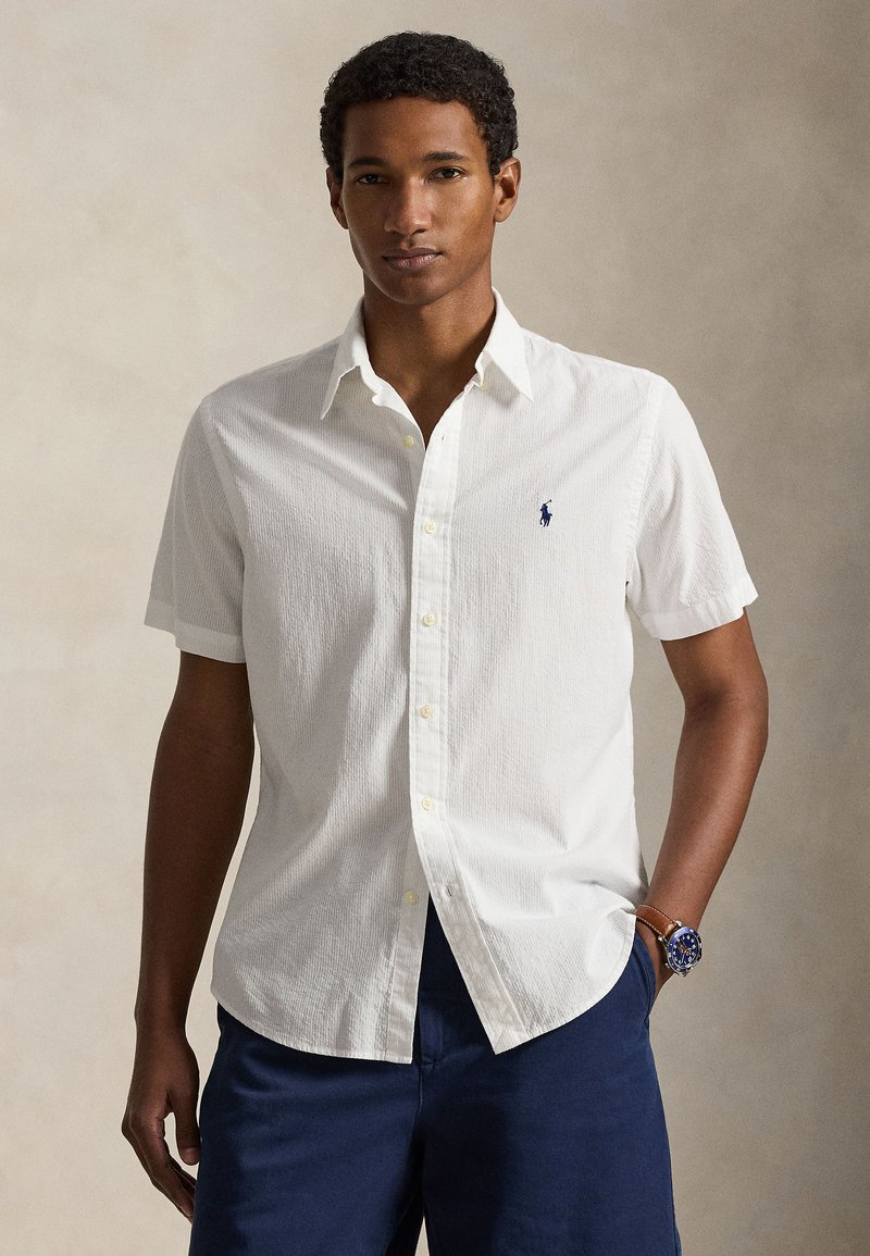 White short-sleeve button-up shirt with a textured fabric, featuring a small blue logo on the chest and yellow buttons. Navy shorts visible.