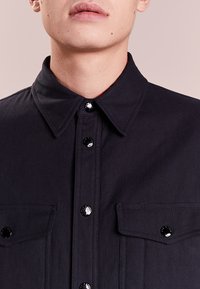Camicia nera con bottoni a scomparsa, dotata di due tasche sul petto, bottoni neri lucidi e colletto a punta. Realizzata in tessuto testurizzato con un design strutturato.