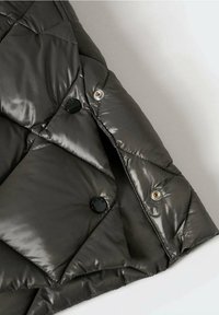 Gros plan sur la manche d'une veste noire matelassée en tissu brillant avec deux boutons-pression noirs, l'un fermé et l'autre ouvert.