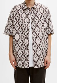 Camisa de manga corta abotonada en marrón oscuro con un patrón floral geométrico blanco, que presenta un corte relajado y botones visibles en el frente.