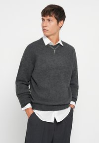 Koolgrijze gebreide trui met geribde manchetten en onderrand, gedragen over een witte blouse met kraag. Zilveren ketting met een hangertje is zichtbaar.