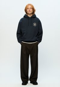 Navy blue hoodie with a logo on the chest, paired with brown pleated trousers and black sneakers. Background is plain white.