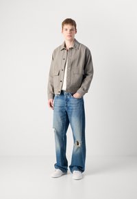 Lys grå knapp-up jakke med to frontlommer, brukt over en hvit skjorte. Slitte blå jeans med store hull. Hvite sneakers.