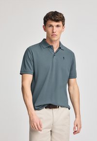 Camisa polo en azul teal, de manga corta, con tapeta de tres botones, que presenta un pequeño logotipo oscuro en el pecho, combinada con pantalones de color claro.