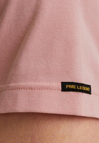 Svetlo roza majica s gladko teksturo, z črno nalepko z rumenim besedilom "PME LEGEND" blizu roba.