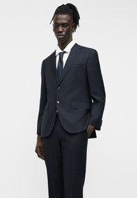 Jeune homme portant un costume bleu marine foncé avec une chemise blanche et une cravate bleue marine, debout avec une main dans la poche devant un fond uni.