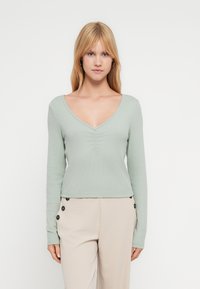 Νεαρή γυναίκα με ίσια ξανθά μαλλιά που φοράει ανοιχτό πράσινο μακρυμάνικο τοπ με V λαιμόκοψη και μπεζ παντελόνι με μαύρα κουμπιά.