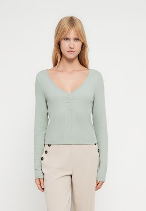 Młoda kobieta o prostych blond włosach, ubrana w jasnozielony, długorękawowy top z dekoltem w serek oraz beżowe spodnie z czarnymi guzikami.