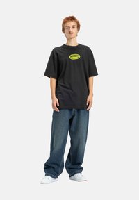 Młoda osoba z krótkimi, jasnobrązowymi włosami nosi dużą czarną koszulkę z żółtym logo, luźne niebieskie dżinsy i białe sneakersy, stoi.