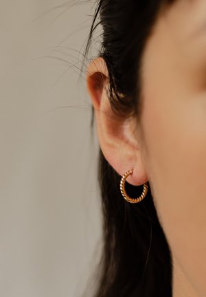 Gold-toned hoop earrings with a twisted design, small in size, worn in a pierced ear against dark hair. Subtle shine and textured finish.